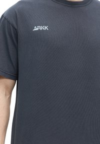 Σκούρο γκρι κοντομάνικο μπλουζάκι με μικρό λευκό λογότυπο "ARKK" στο πάνω αριστερό μέρος του στήθους, φορεμένο από άτομο με μερικώς ορατό χέρι.