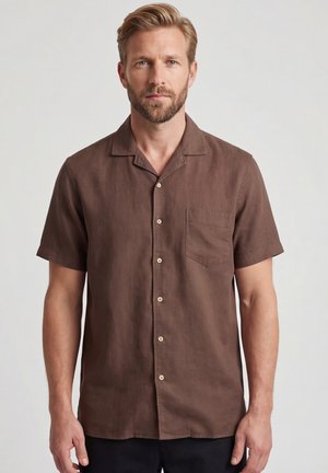 Homme blond avec une barbe portant une chemise marron à manches courtes avec une poche poitrine, debout devant un fond clair uni.