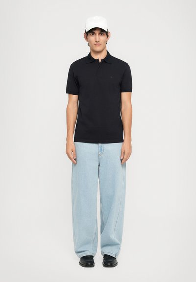 Man som står mot en vit bakgrund iförd en svart polo, ljusblå vida jeans, svarta skor och en vit basebollkeps.