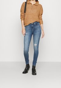 Pull en maille marron avec col, associé à un jean skinny bleu avec des détails usés. Porté avec des bottines noires.