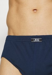 Námořnické modré bavlněné trenkoslipy s jemnou texturou, mají elastický pas a malou štítkovou nášivku. Detailní záběr na látku a barvu.