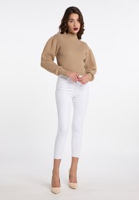 Maglione beige a coste con collo alto e maniche a sbuffo, abbinato a jeans skinny bianchi e décolleté nudi, su uno sfondo neutro.