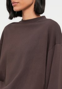 Καφέ, υπερμεγέθη φούτερ με ψηλό λαιμό και πέφτουσες άκρες στους ώμους. Μαλακό ύφασμα με λείο υφή, μινιμαλιστικός σχεδιασμός, χωρίς σχέδια.