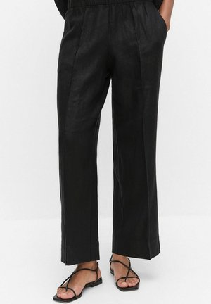 Zwarte wijde broek gedragen door een persoon met handen in de zakken, gecombineerd met zwarte platte sandalen met bandjes tegen een effen witte achtergrond.