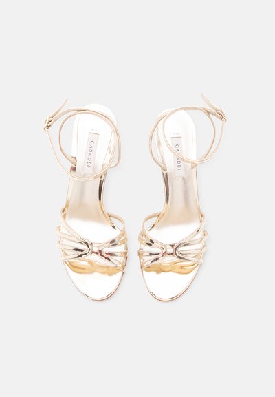 Ζευγάρι χρυσά σανδάλια με λουριά και αγκράφες στον αστράγαλο, ανοιχτά δάχτυλα και επίπεδες σόλες, εμφανιζόμενα από πάνω σε λευκό φόντο.