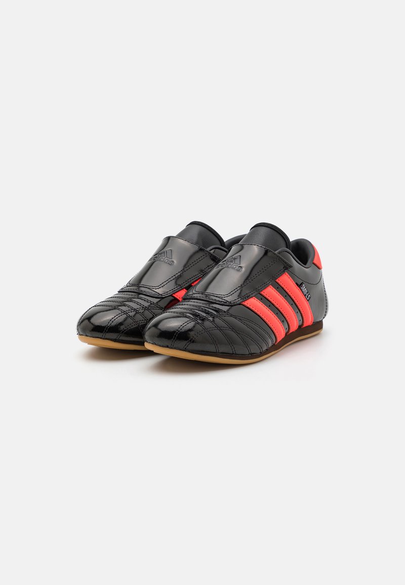 Zapatillas deportivas de cuero negro con acentos rojos. Incluyen características como una punta redondeada, parte superior texturizada y suela de goma, mostrando tres franjas laterales.