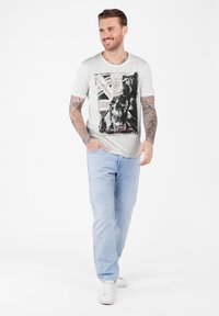 Hombre con brazos tatuados que lleva jeans claros, zapatillas deportivas blancas y una camiseta blanca con un gráfico de calavera y la palabra "Libertad", caminando y sonriendo.