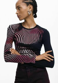 Czarna bluzka z długim rękawem o teksturowanym, wielokolorowym wzorze w fale w odcieniach różu i bieli. Dopasowany krój z prążkowanej tkaniny. Modelka pozowała z rękami skrzyżowanymi.