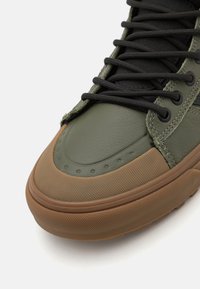 Grønn lærersneaker med teksturert brun gummisåle, rund tå, svarte snøringer og perforerte detaljer på skoens vamp.