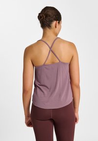 Débardeur de sport violet avec un ourlet arrondi et des bretelles croisées dans le dos, fabriqué en matière douce et respirante. Porté avec des leggings bordeaux.