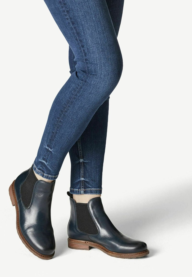 Personne portant des bottines Chelsea en cuir bleu foncé avec des semelles marron et un jean skinny bleu foncé sur un fond uni.
