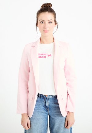 Ung kvinde med hår sat op i knold iført pink blazer, hvid t-shirt med teksten "HAPPY MOOD" og solgrafik samt blå jeans, stående foran en hvid baggrund.