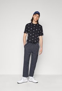 Black t-shirt with repeating purple Nike logos, paired with gray, loose-fit pants featuring an adjustable drawstring and a pocket. White sneakers.