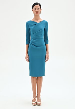 Femme debout, portant une robe sarcelle mi-longue avec des manches trois-quarts et des détails drapés, des boucles d'oreilles créoles dorées et des talons dorés à brides.