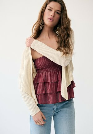 Jeune femme portant un cardigan en maille crème sur un haut maroon sans bretelles à volants, associé à un jean bleu clair et de grandes boucles d'oreilles géométriques.