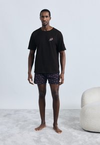 Tricou negru din bumbac cu broderie de cireșe, asortat cu pantaloni scurți închiși la culoare cu un model de fructe roșii, purtat cu picioarele goale pe un covor gri deschis.