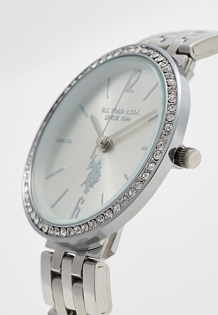 Montre en acier inoxydable argenté avec un cadran orné de cristaux, un cadran argent clair et des index en contraste. Elle est dotée d'un design de bracelet classique.