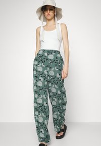 Calças largas com padrão floral em verde, adornadas com flores brancas e folhas pretas, combinadas com uma t-shirt branca e um chapéu de sol cinza. Sandálias pretas.