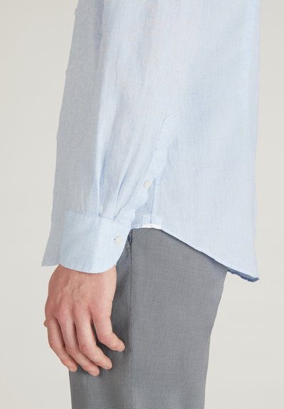 Mano maschile appoggiata di lato, indossando una camicia azzurro chiaro a maniche lunghe con bottoni e pantaloni grigi texturizzati, su uno sfondo neutro.