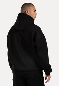 Zwarte hoodie met een gladde textuur en een oversized pasvorm. Voorzien van een grote capuchon en geribbelde manchetten, ontworpen voor comfort en warmte.