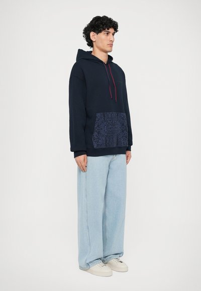 Marineblaue Kapuzen sweatshirt mit ropem Kordelzug und einer gemusterten marineblauen Tasche, kombiniert mit hellblauen weiten Jeans und beigen Sneakers.