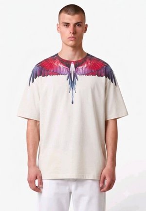 Uomo con capelli corti che indossa una maglietta crema con design di ali di uccello rosse, viola e blu sulle spalle e sul petto.