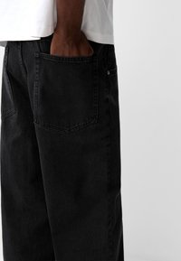 Pantalones de mezclilla negra con un corte relajado, que cuentan con un bolsillo trasero visible y detalles de costura. La textura es suave con un ligero brillo.