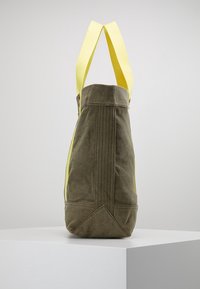 Canvas shopperväska med olivgrön kropp, ljusgula remmar och vertikala sömmar. Botten har en vinklad, strukturerad form.