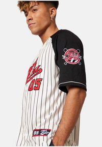Tricou de baseball cu mâneci negre și corp alb cu dungi fine. Accente roșii pe față și logo-ul de pe mânecă, cu țesătură texturată.