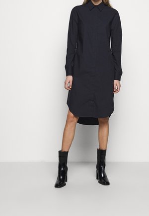 Kobieta ubrana w czarną sukienkę koszulową z długim rękawem i czarne botki na obcasie, stojąca na tle jednolitego białego tła.