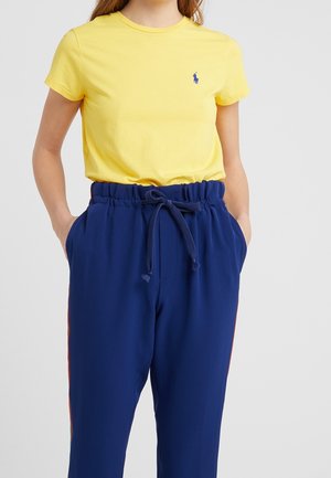 Osoba ubrana w żółtą koszulkę z krótkim rękawem z niebieskim logo oraz granatowe spodnie z sznurkiem w pasie, z rękami w kieszeniach.