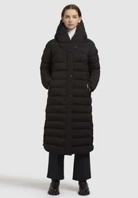 Een vrouw in een lang zwart pufferjasje met een grote kraag, zwarte broek en zwarte laarzen, staand tegen een eenvoudige lichte achtergrond.