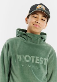 Felpa verde in pile con logo "PROTEST" in rilievo e colletto alto. Indossata con un cappellino nero e beige decorato con un emblema.