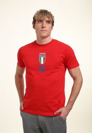 Giovane uomo in piedi con le mani nelle tasche, che indossa una maglietta rossa con lo stemma dell’Italia, quattro stelle e il numero 82 sul davanti.