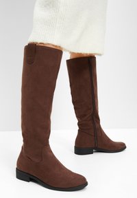 Stivali al ginocchio in suede marrone con zip laterale, suola piatta e punta arrotondata. Texture liscia con dettagli decorativi minimi.