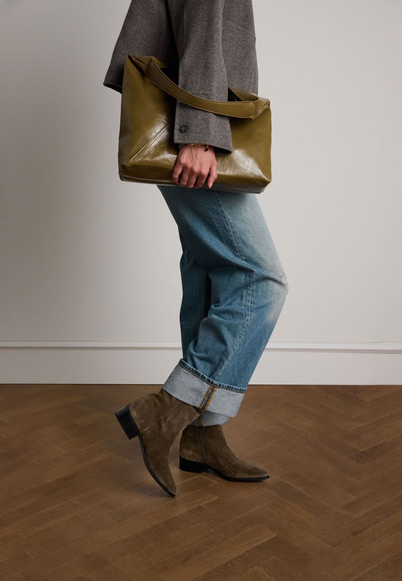 Borsa in pelle verde oliva con design strutturato, abbinata a jeans chiari con risvolto e stivaletti in suede marrone su un pavimento di legno.