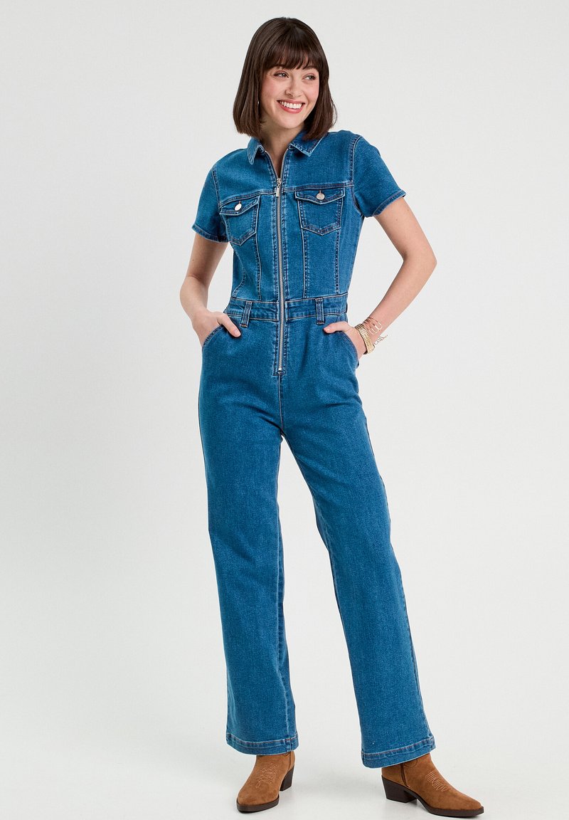 Salopette in denim con maniche corte, zip frontale, due tasche sul petto e vita aderente. Il colore è blu medio con un tessuto texturizzato.