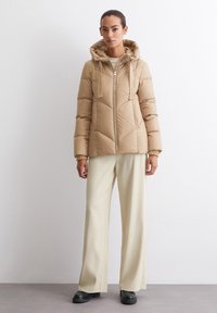Beige puffer jacket with a hood and zip front, featuring a quilted design. Paired with cream wide-leg trousers and black footwear.