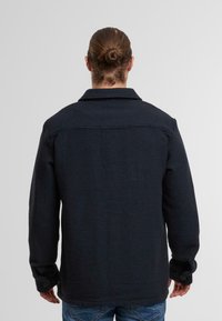 Marineblaues gewebtes Jacket mit strukturierter Oberfläche, geknöpften Manschetten und spitzem Kragen. Verfügt über ein Yoke-Design auf der Rückseite.