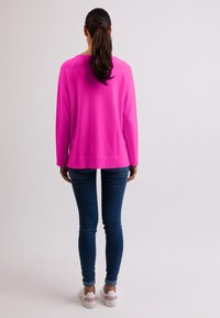 Fuchsiový svetr s dlouhými rukávy, kulatým výstřihem a žebrovanými manžetami, spárovaný s tmavě modrými skinny džíny a bílými teniskami.