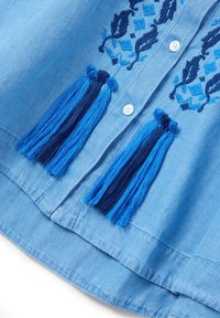Camicia azzurra con motivi ricamati in blu scuro, caratterizzata da due lunghe frange blu e bottoni bianchi. Tessuto morbido e liscio.