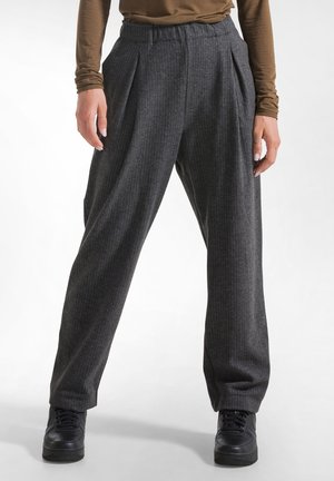 Persoon draagt hoog getailleerde donkergrijze pinstripe wijde broek met plooien en zwarte sneakers tegen een effen lichte achtergrond.