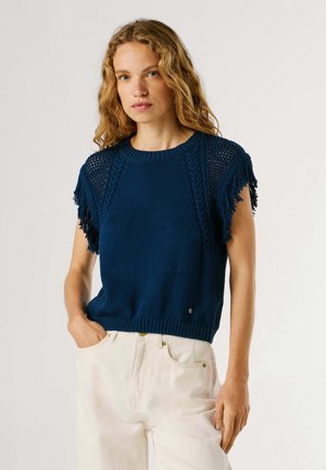 Donna con capelli biondi mossi che indossa un top senza maniche in maglia blu navy con spalle sfrangiate e pantaloni bianchi, in piedi davanti a uno sfondo semplice.