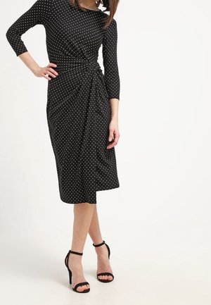 Frau trägt ein schwarzes Midikleid mit weißen Polka Dots und seitlicher Knotendetail, kombiniert mit schwarzen High Heels mit Knöchelriemen.