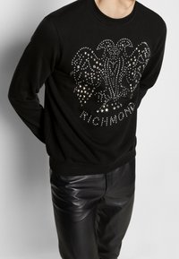 Svart sweatshirt i mjukt tyg, med en nitar design som har ett dekorativt emblem och ordet "RICHMOND" i silverdetaljer.