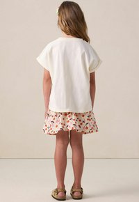 Un haut en coton blanc à manches courtes, associé à une jupe évasée ornée d'un motif de cerises en orange et rose. Des sandales beige complètent la tenue.