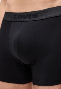 Κοντινό πλάνο ενός ατόμου που φοράει μαύρα ανδρικά εσώρουχα Levi's boxer briefs με ορατό λογότυπο στη μέση και λεία υφή υφάσματος.
