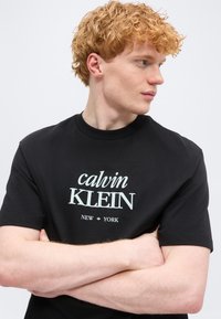 Νέος άνδρας με σγουρά κόκκινα μαλλιά, φοράει μαύρο μπλουζάκι Calvin Klein New York, με σταυρωμένα τα χέρια, κοιτάζει προς τα δεξιά του.