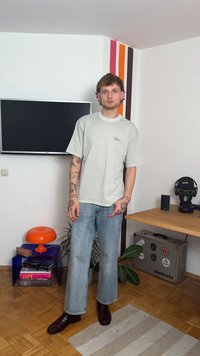 Ανοιχτό γκρι oversized μπλουζάκι με λογότυπο, μπλε φαρδιά τζιν παντελόνια, σκούρα καφέ παπούτσια, ριγέ υφασμάτινο χαλί με κουκίδες, μοντέρνα έπιπλα.
