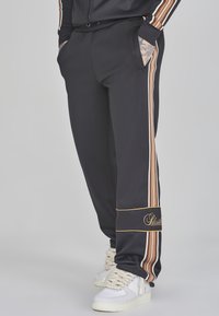 Zwarte sportbroeken met verticale strepen in oranje, wit en bruin, met zijzakken en een gouden geborduurd logo bij de zoom.